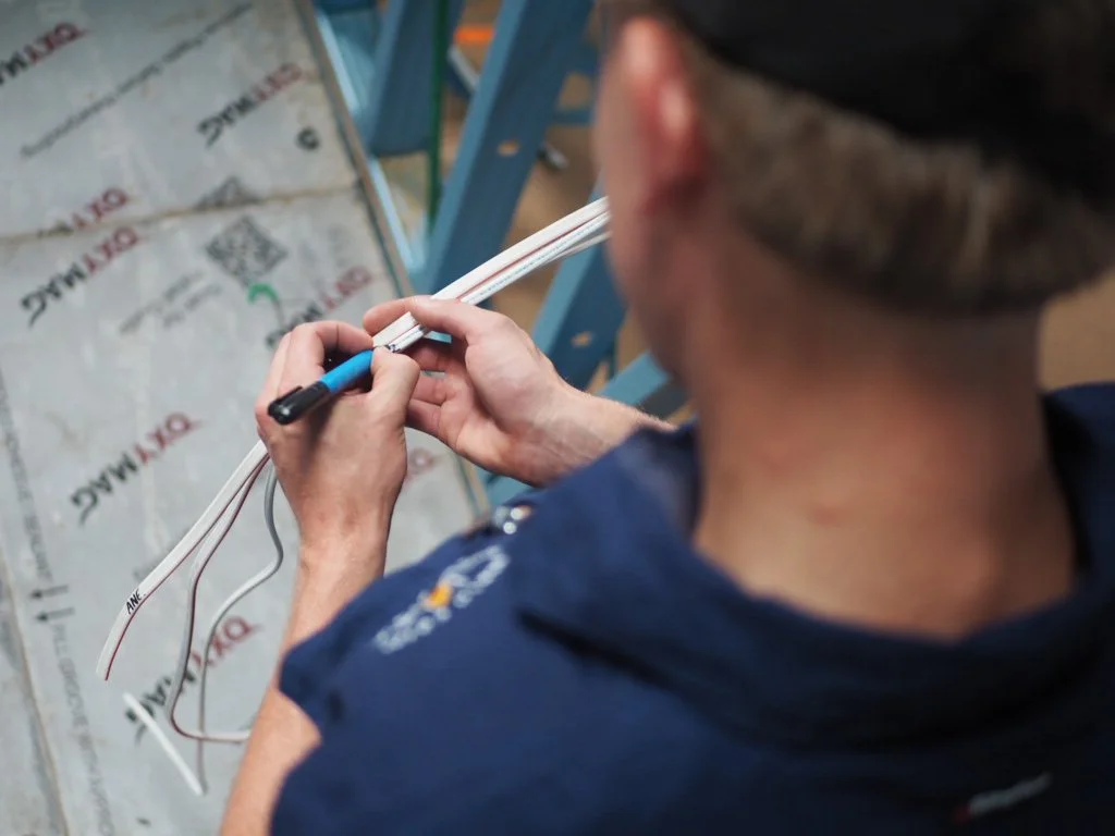 A person working on electrical wiring with a marker, close to a building's insulation material and blue support beams.
