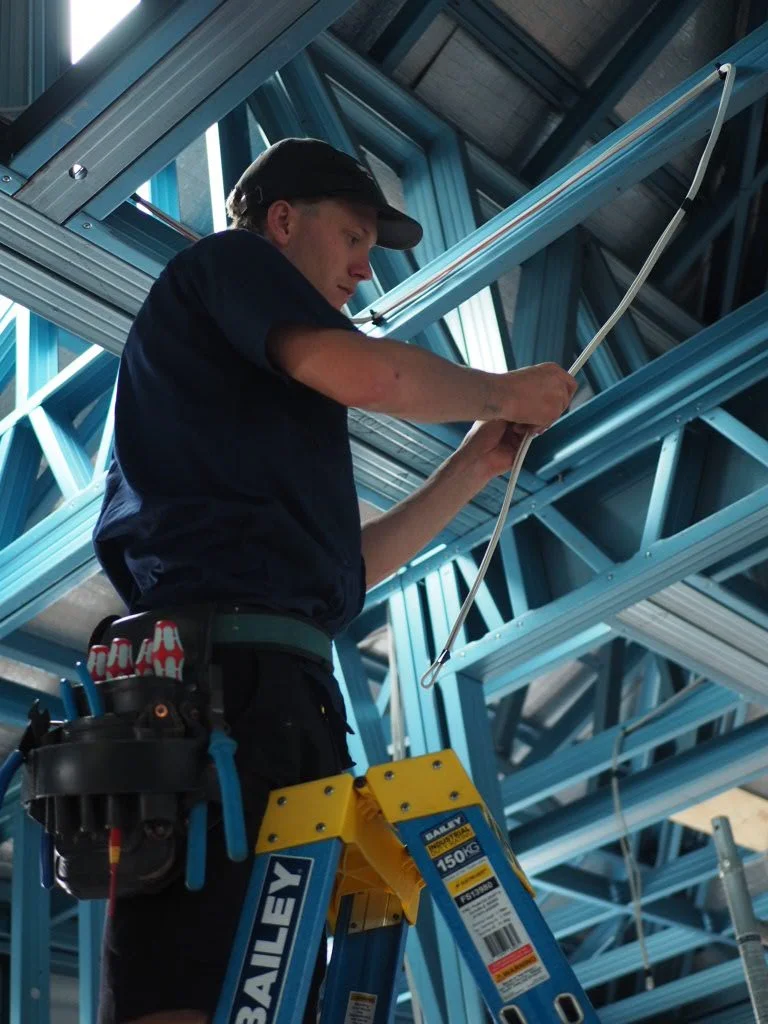 A young male construction worker on a yellow ladder installing or inspecting blue metal framing in a building under construction.