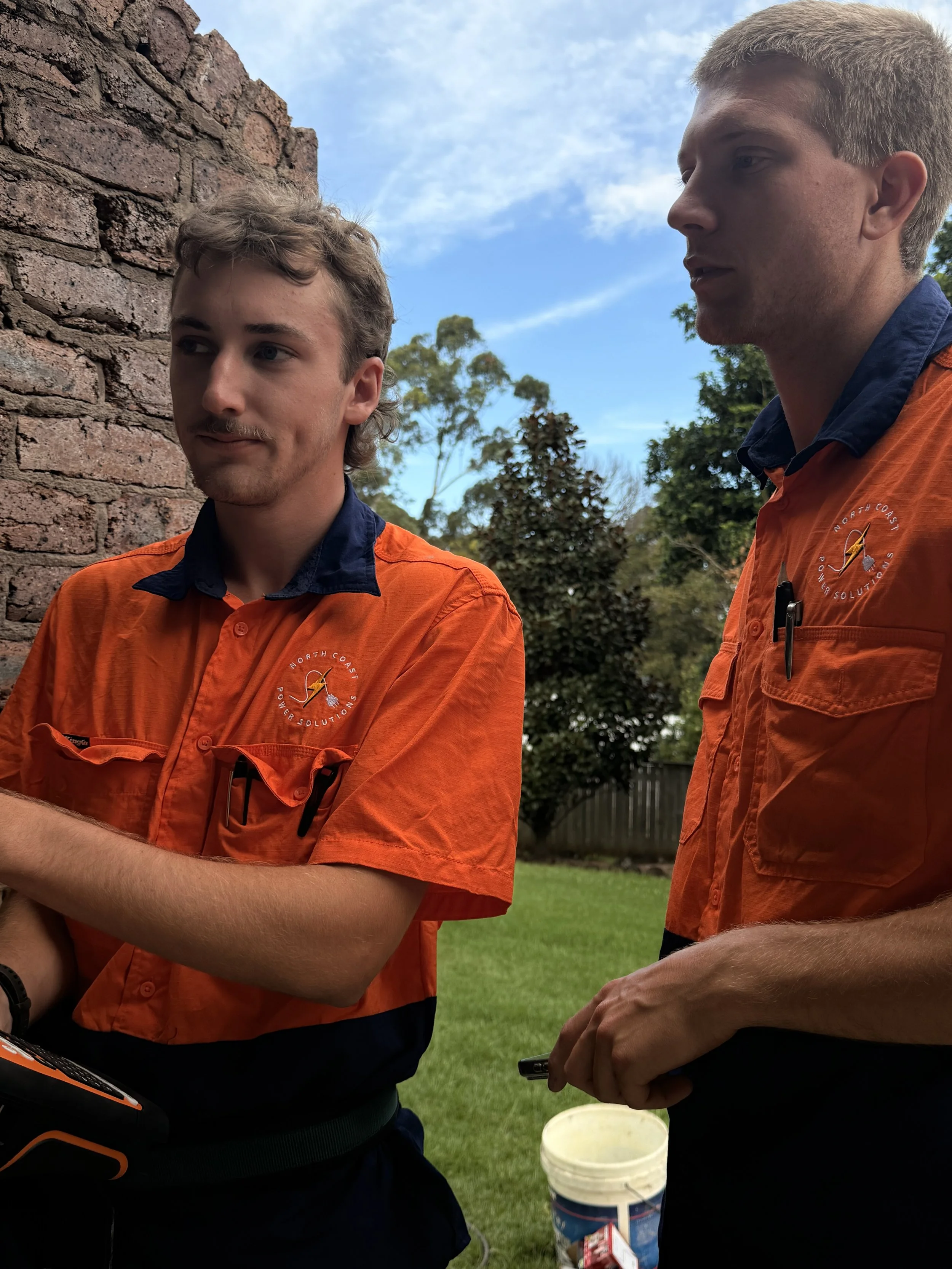 Two young men wearing matching orange work uniforms with a logo on the chest, standing outdoors near a brick wall and trees, with a cloudy sky in the background.