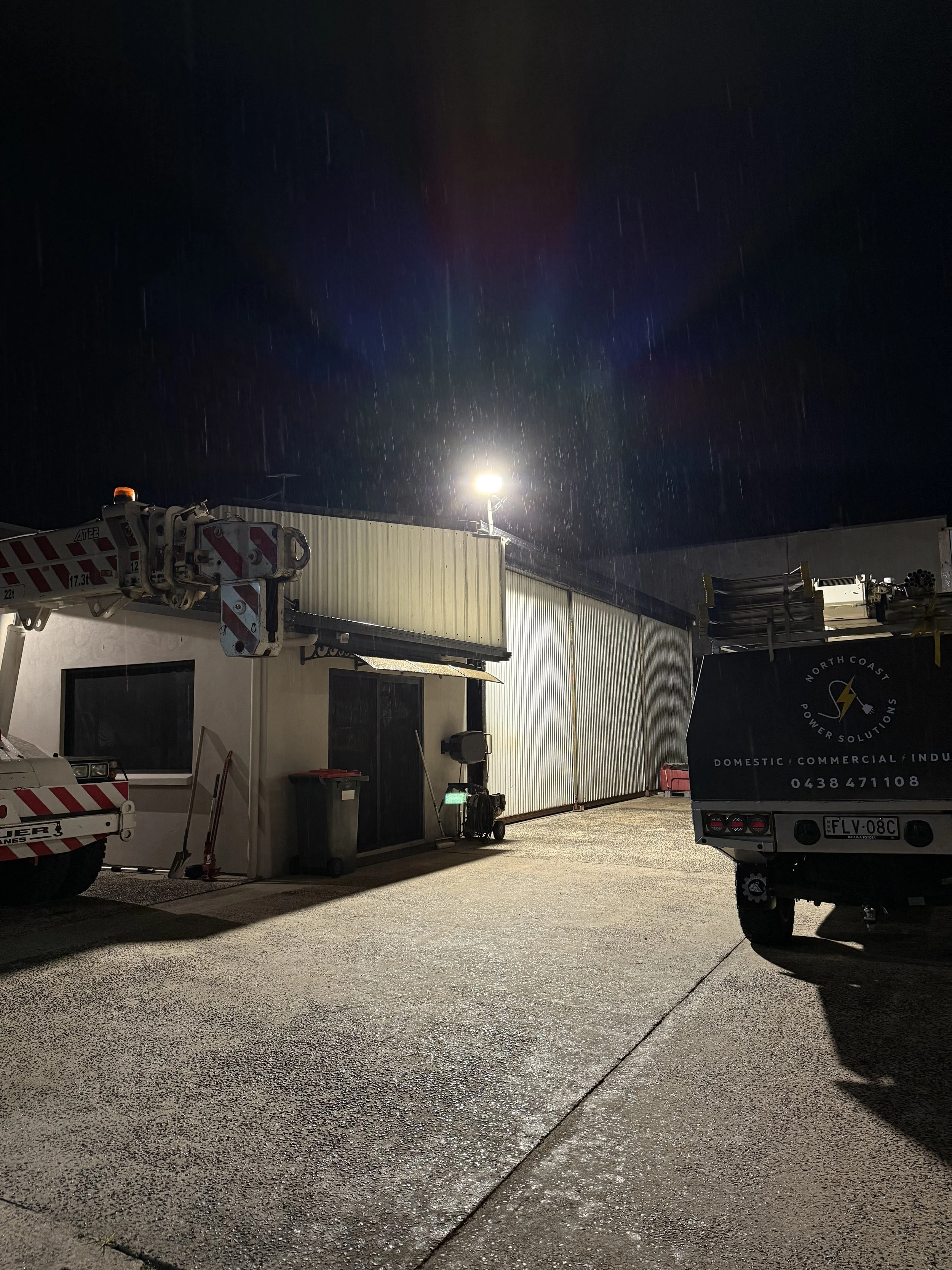 Nighttime scene of an industrial or commercial area with a building, vehicles, and equipment illuminated by bright lights. Rain is falling, and the ground appears wet.
