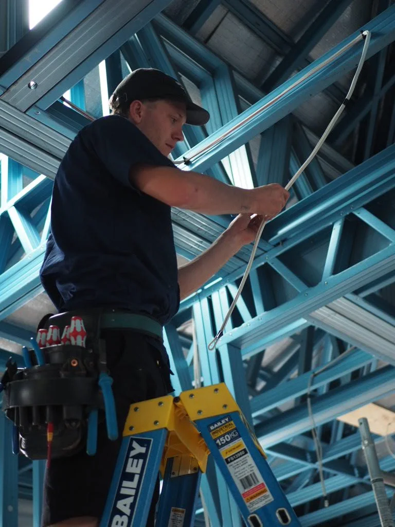 A construction worker standing on a yellow ladder, installing or inspecting electrical wiring in a steel-framed building.