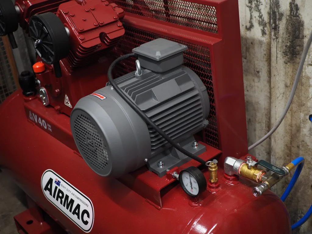 Close-up of a red AIRMAC portable air compressor with a gray motor, pressure gauge, and brass fittings, positioned against a textured wall.