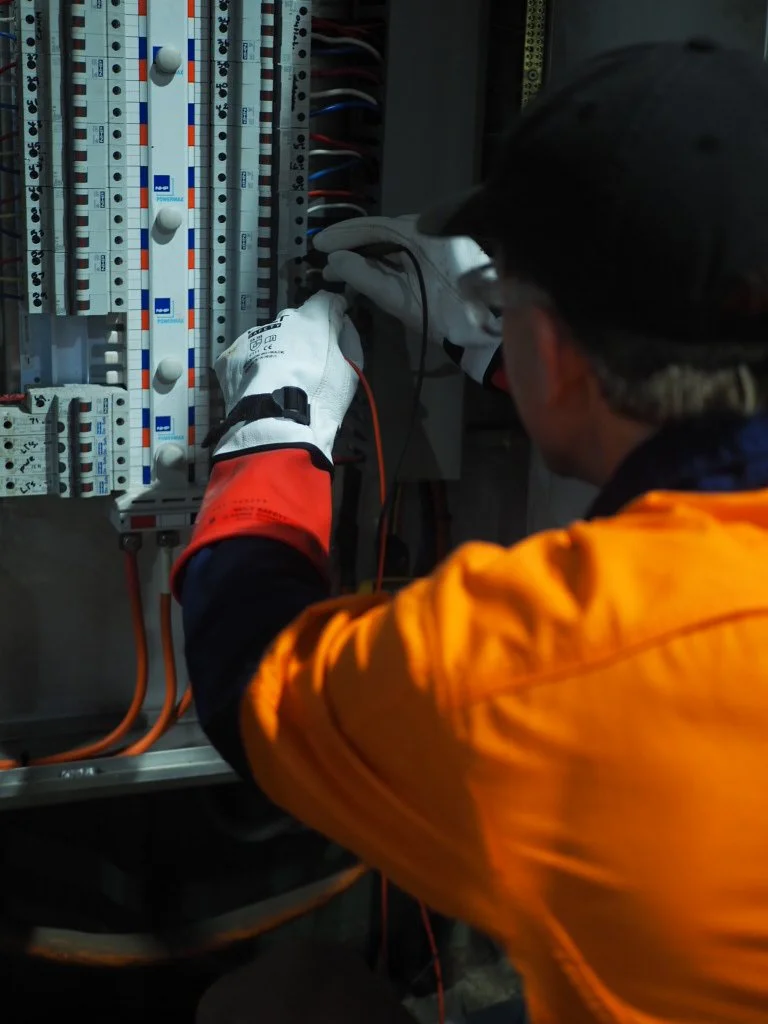 Worker in safety gear working inside an electrical panel with switches and wiring.