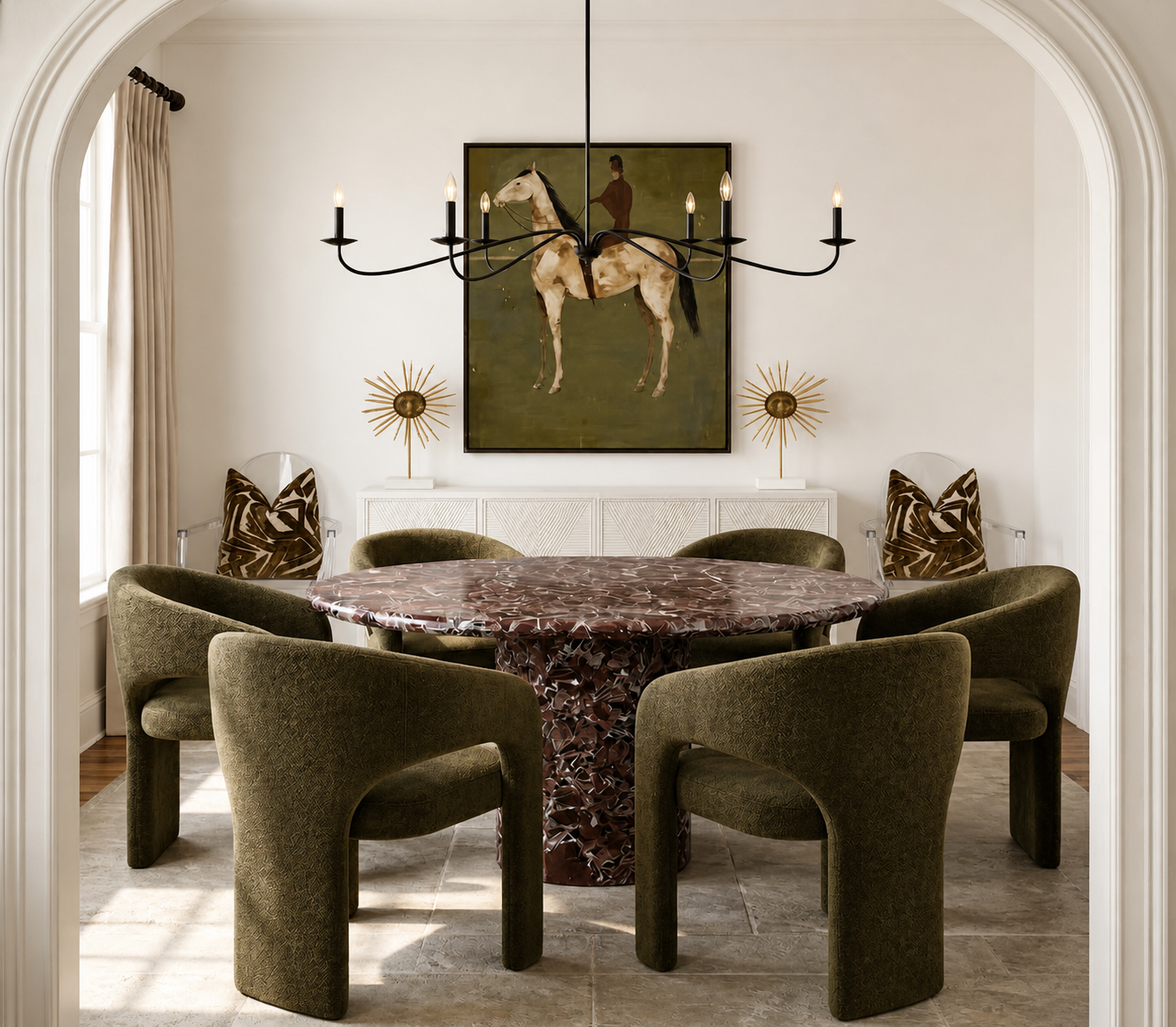 A dining room with a round table, six green velvet chairs, two armchairs with patterned pillows, a chandelier, art, and decorative objects.