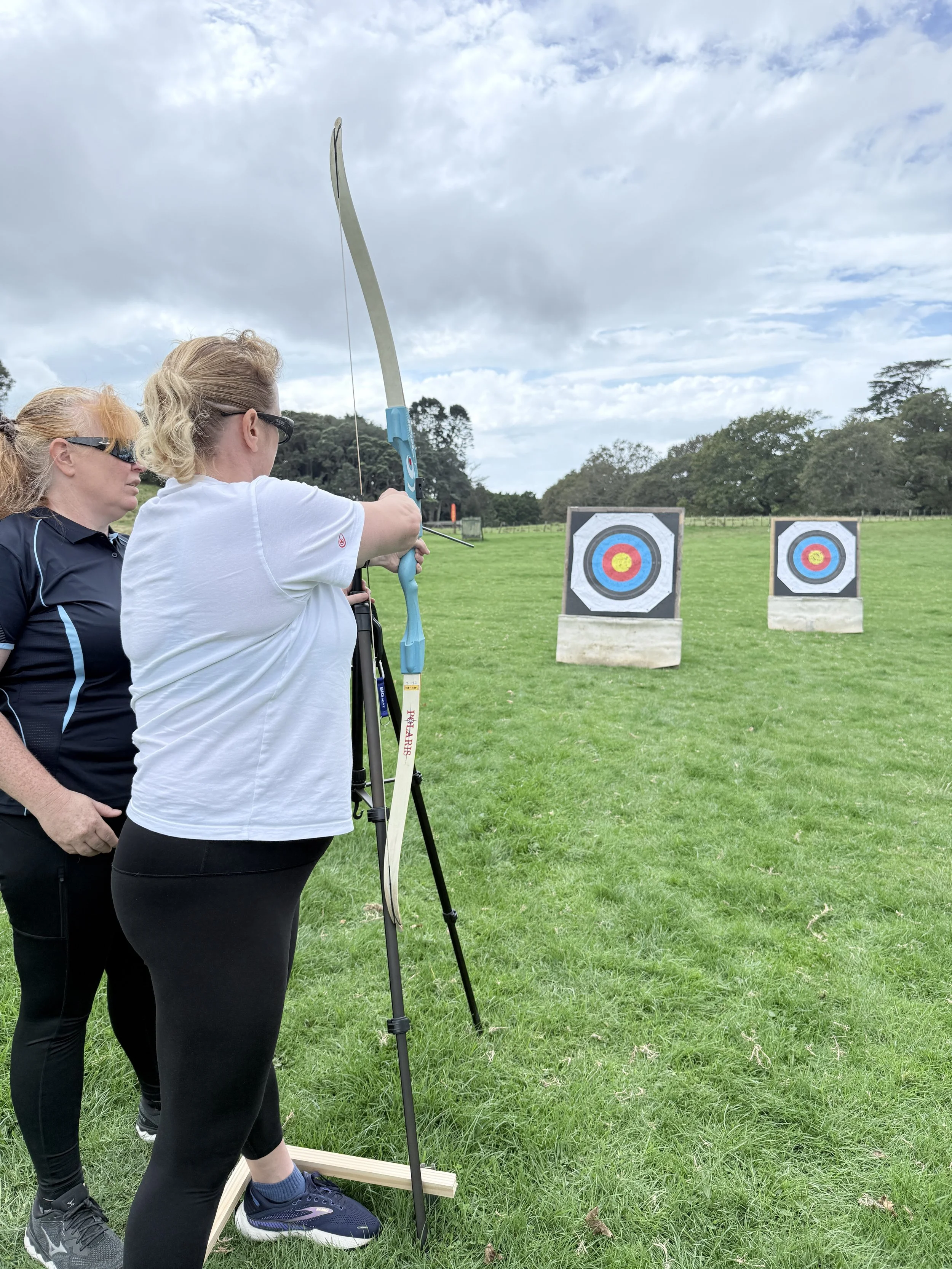 Two women are participating in archery outdoors on a grassy field, with two archery targets in the background. The woman in the white shirt is drawing a blue and white bow, aiming at the targets, while the other woman observes.