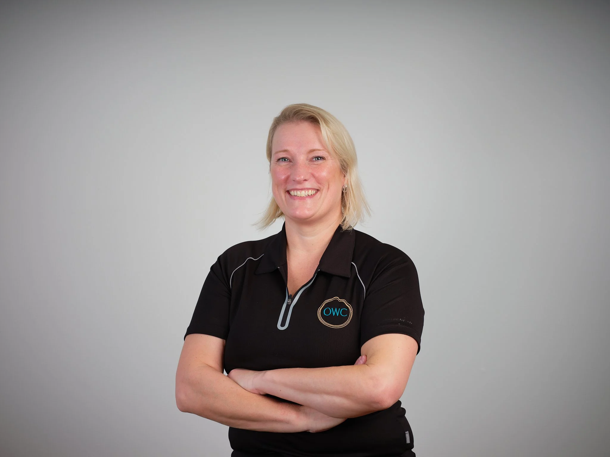 A smiling woman with blonde hair, wearing a black collared shirt with a logo, standing with arms crossed against a plain gray background.