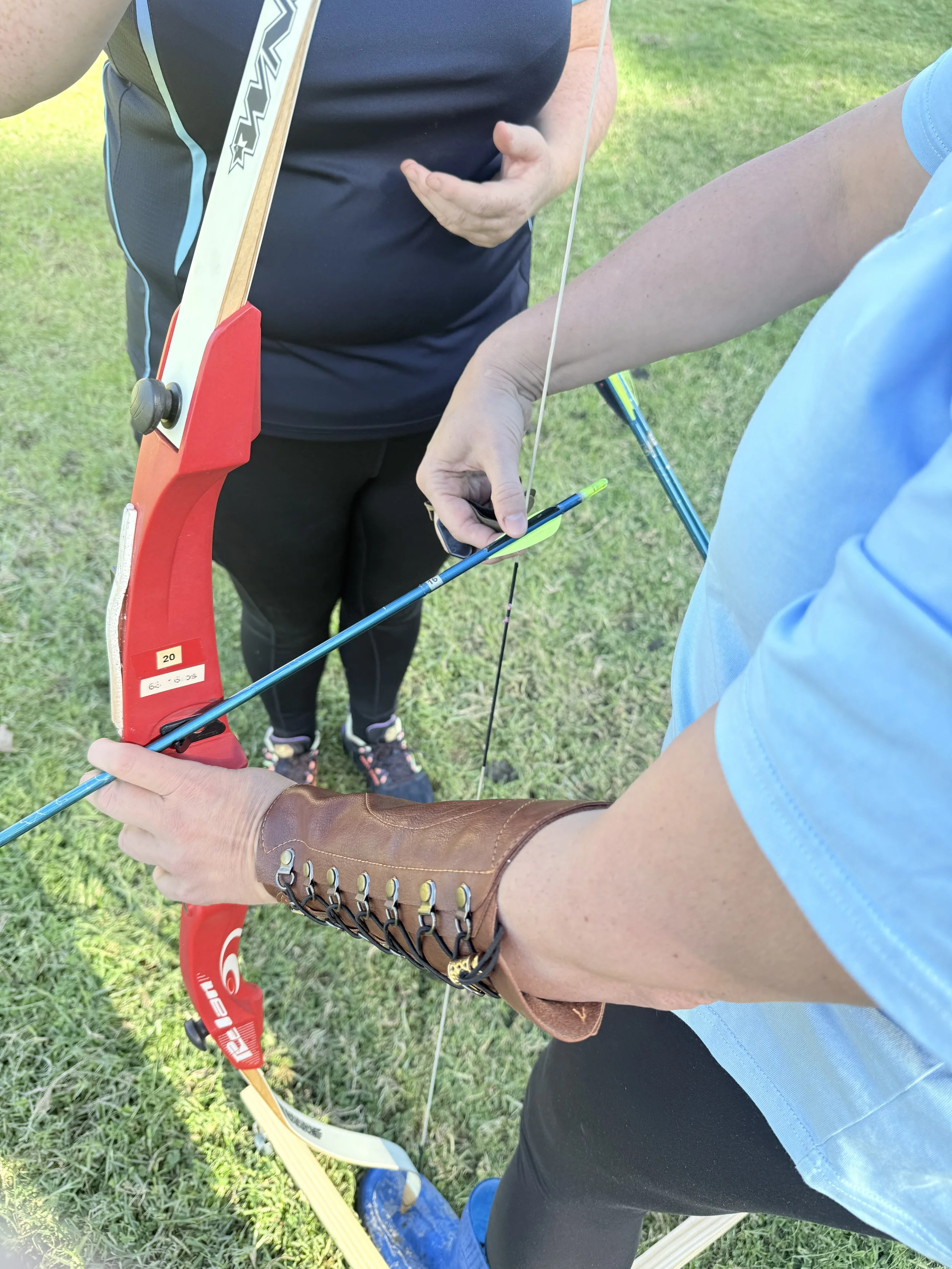 A person wearing a leather arm guard drawing a bow, with another person standing beside them holding an arrow, on a grassy outdoor field.