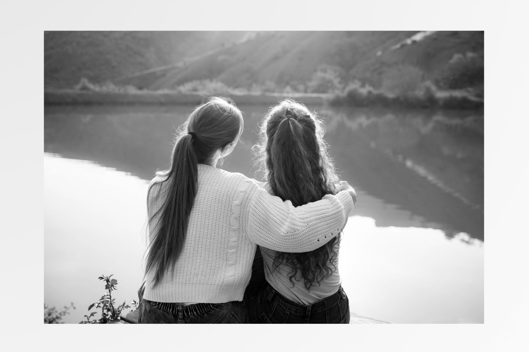 Two women with long hair, one with a ponytail, sit by a lake with mountains in the background, hugging and looking at the water.