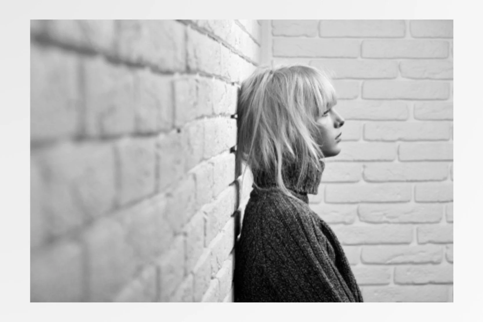 A black and white photo of a woman with shoulder-length hair leaning against a brick wall, looking to the right.