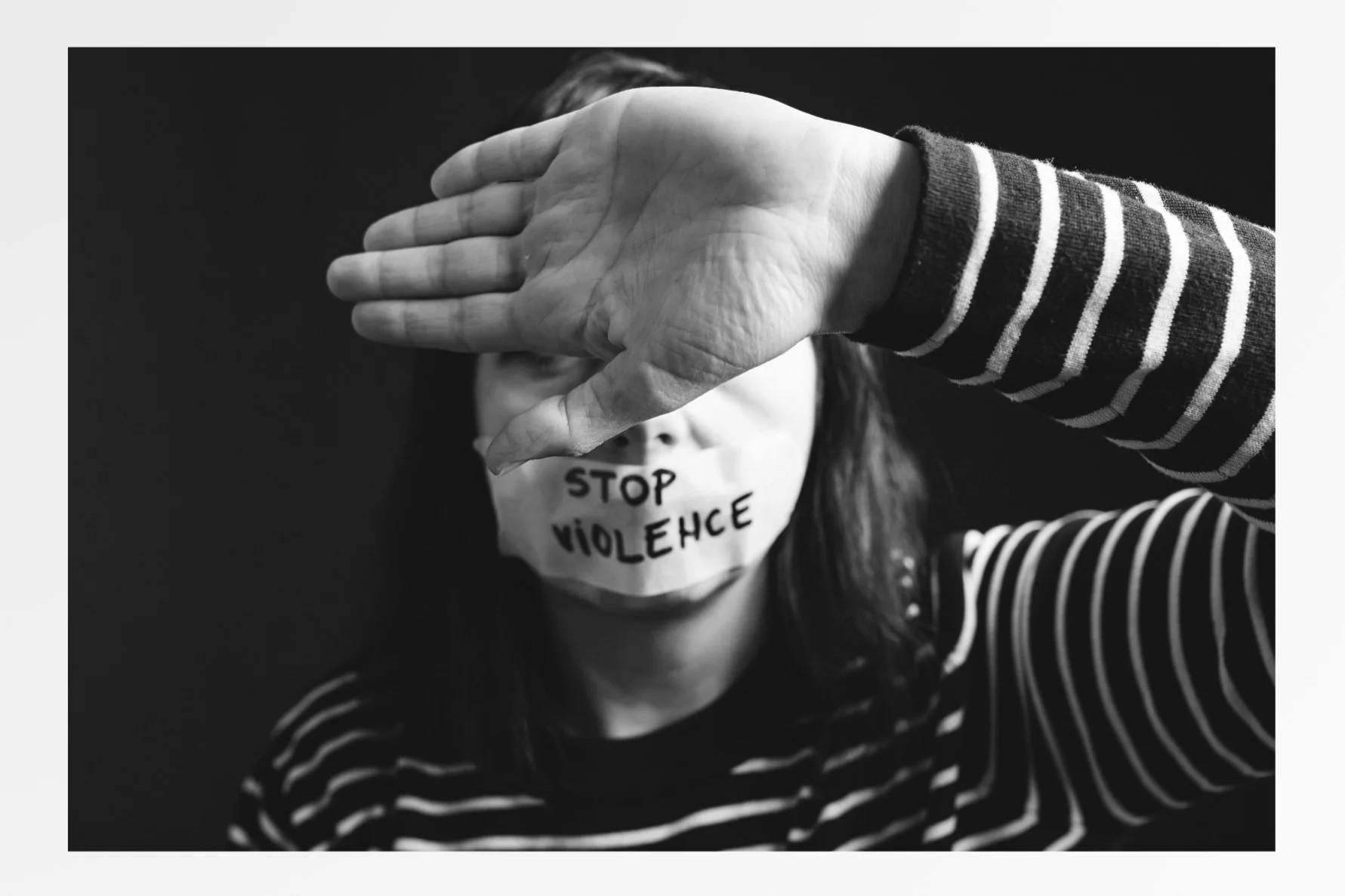 Black and white photo of a woman with a sign over her mouth reading "STOP VIOLENCE". A person with an outstretched hand covers her face, blocking her eyes.