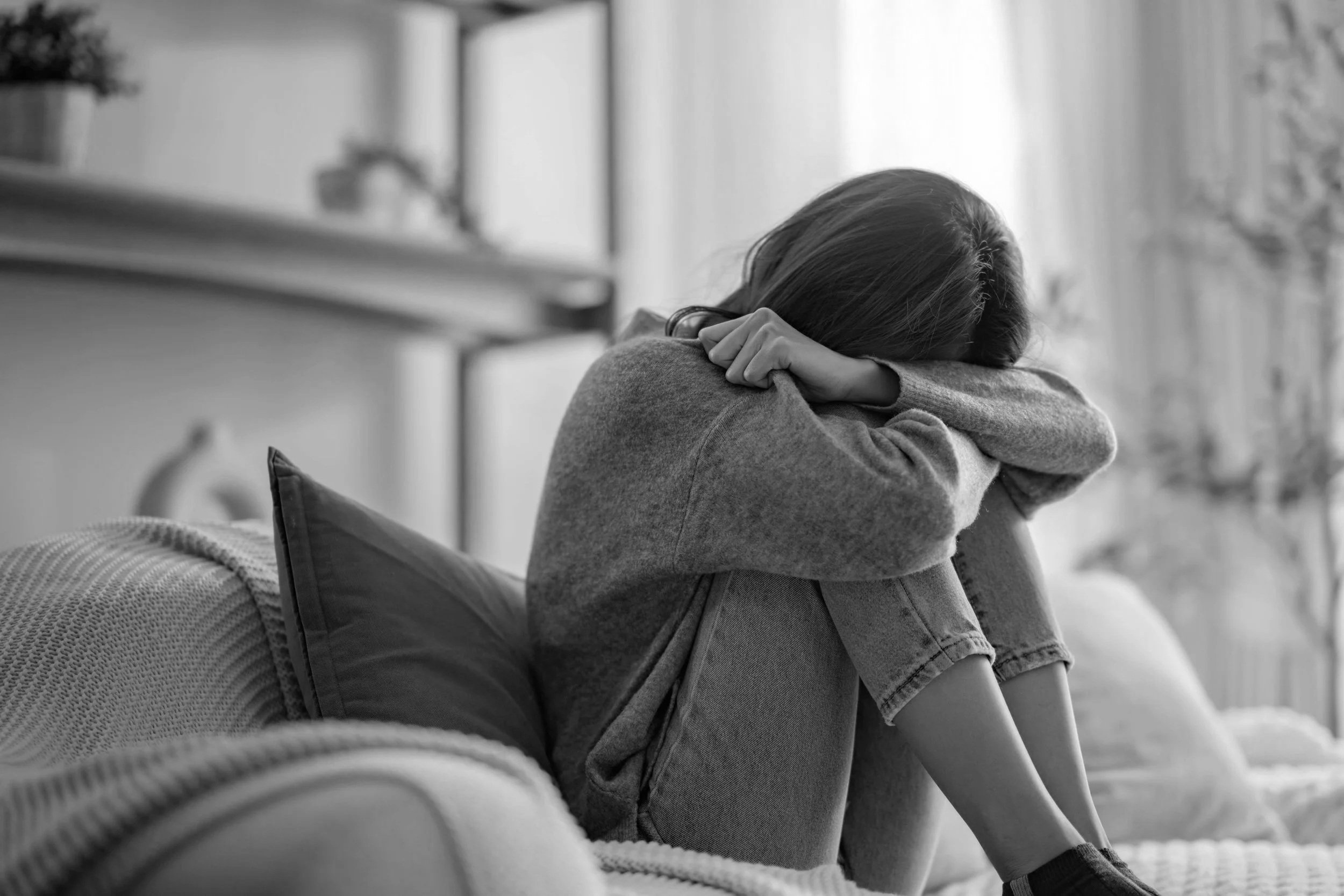 A person sitting on the floor with arms resting on knees and head down, appearing to be sad or distressed in a home setting.