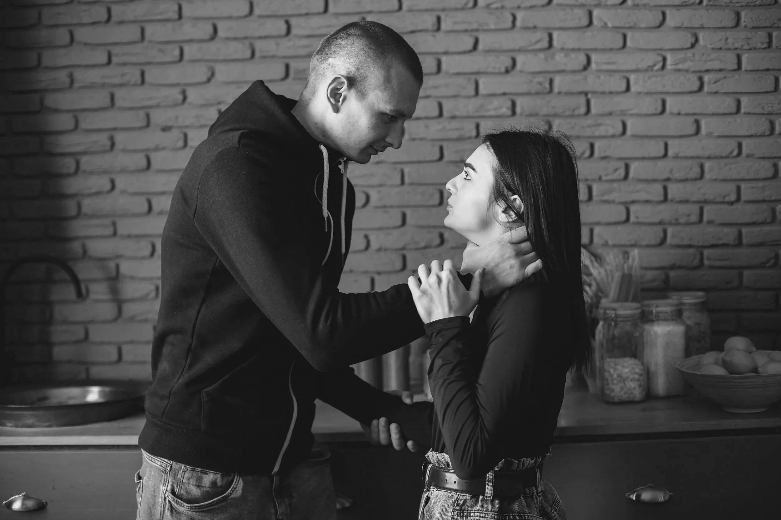 A black-and-white photo of a man and a woman in a kitchen. The man has a shaved head, wearing a black hoodie, and is holding the woman's chin while leaning in close. The woman has long dark hair, wearing a black jacket, and is looking up at the man. The background features a brick wall and kitchen items.