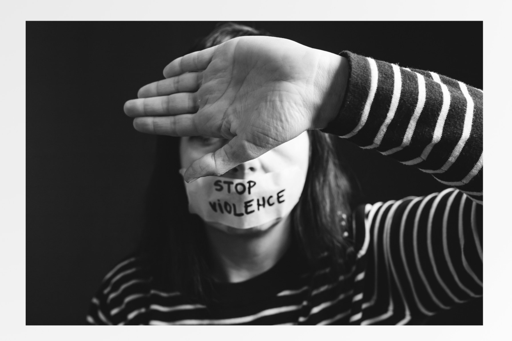 A black and white photo of a woman wearing a striped sweater, with a face mask that says 'STOP VIOLENCE'. She is holding her hand up in front of her face, blocking part of her face and eyes.