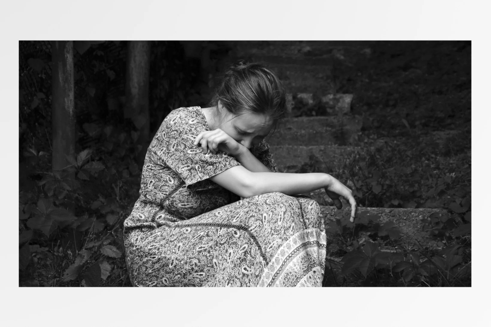 A woman sitting outdoors on a stone or concrete surface, wearing a patterned dress, with her head bowed and covering her mouth with her arm, appearing to be in distress or sorrow.