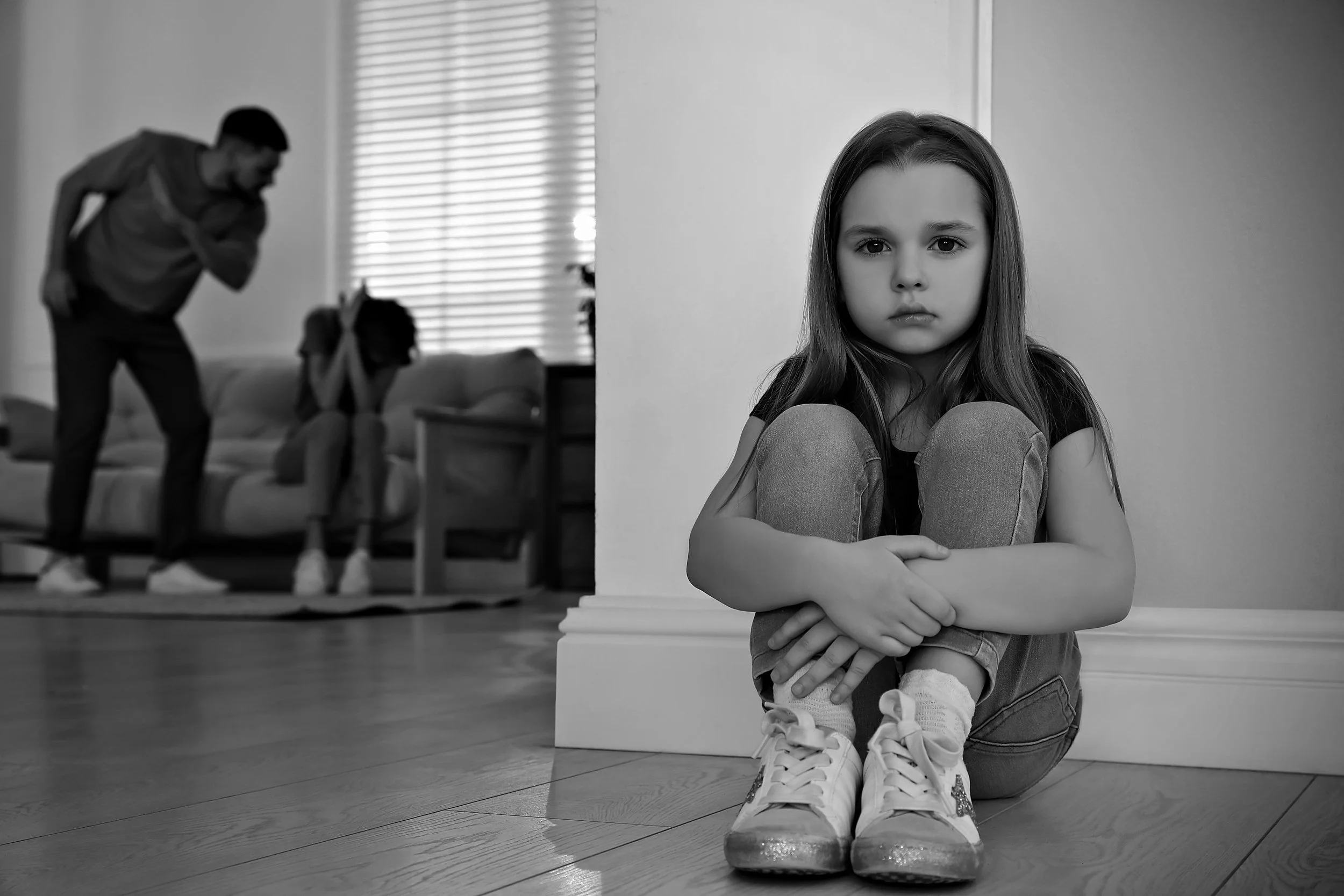 A girl in casual clothes and sneakers sitting on the floor, hugging her knees, looking at the camera, with a serious expression. In the background, a woman and a girl are blurred, sitting on a sofa, with the woman leaning forward and the girl covering her ears. The room has horizontal blinds and wooden flooring.