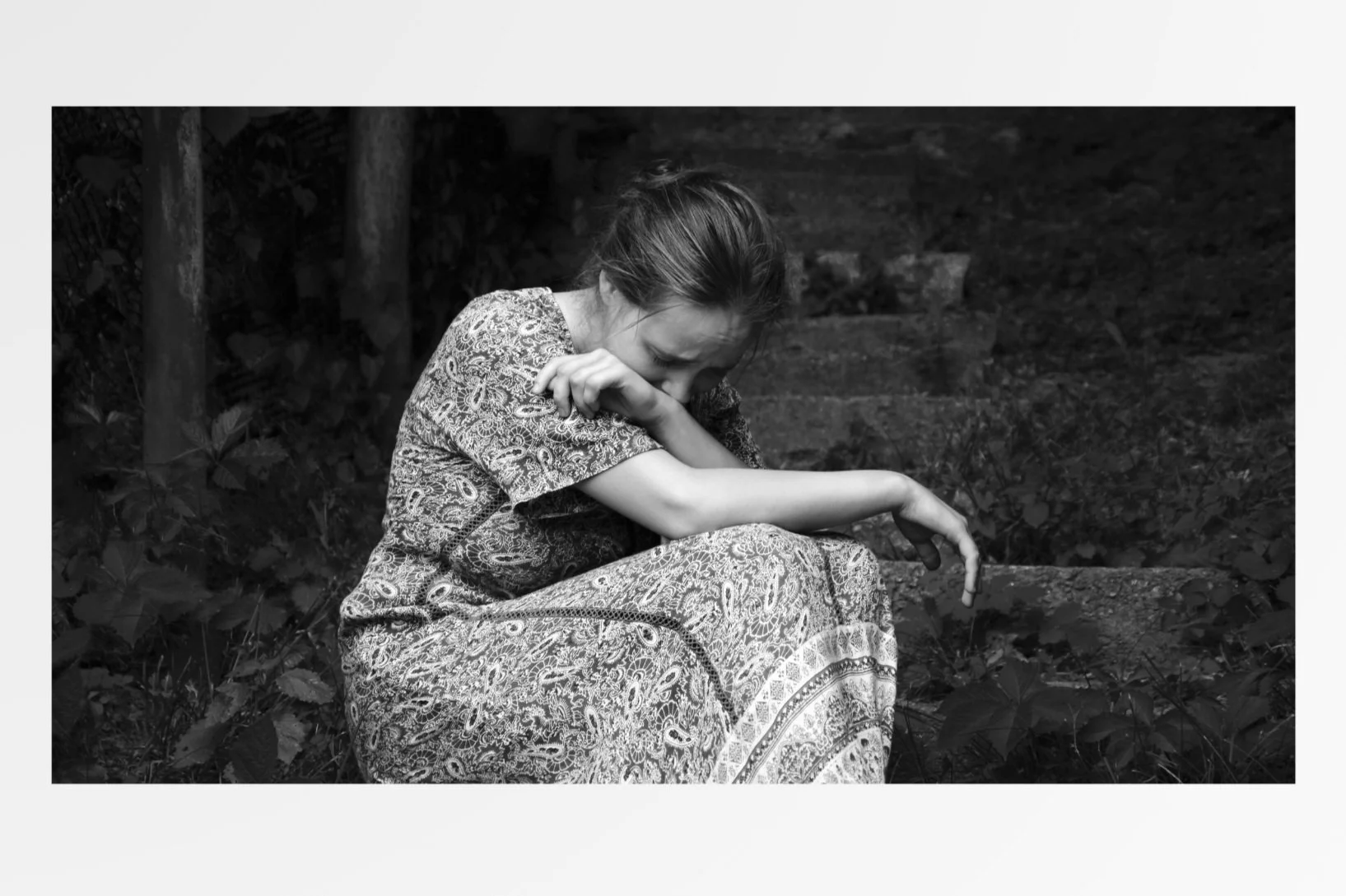 A young girl sitting outdoors on the ground, wearing a patterned dress, appears to be crying or upset, with her head bowed and one arm covering her face.