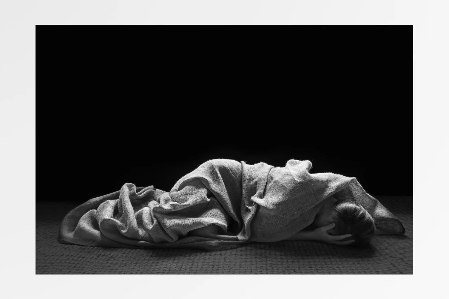 A person lying on the floor, covered with a blanket, resting their head on their arm, in a dimly lit setting.
