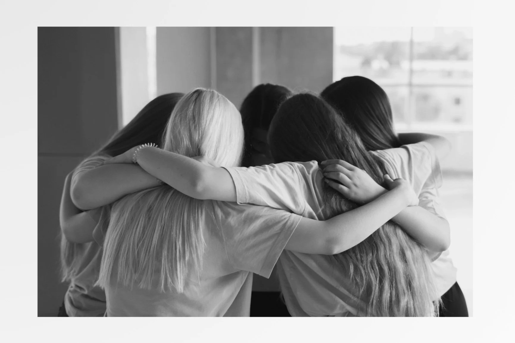 Group of children hugging each other in a close embrace, indoors near a window.