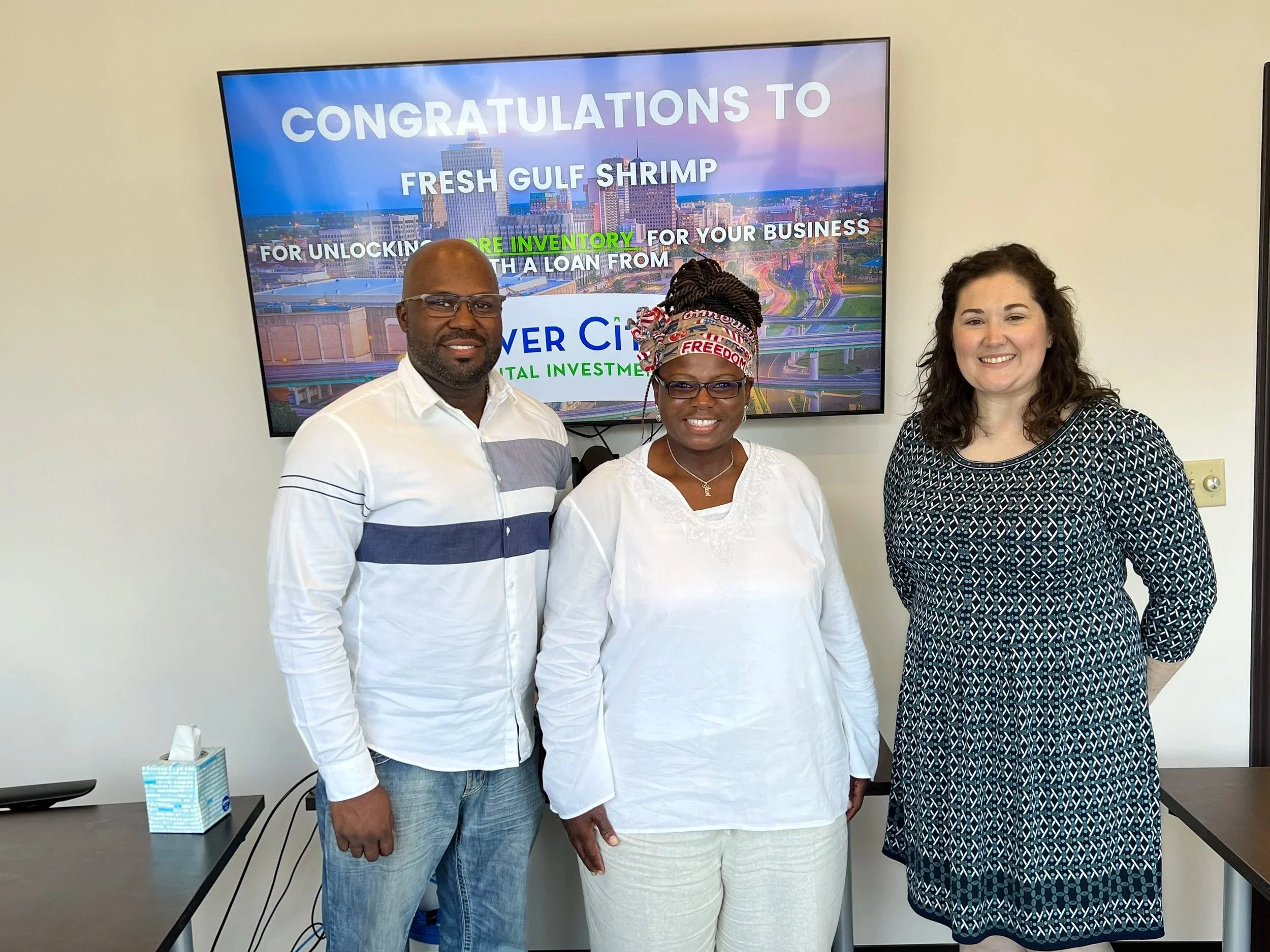 Three people standing in front of a large screen displaying a cityscape and congratulatory text. The man on the left is wearing a white and gray striped shirt and glasses, the woman in the middle is wearing a white blouse and headscarf, and the woman on the right is wearing a black and white patterned dress. They appear to be celebrating or recognizing an achievement.