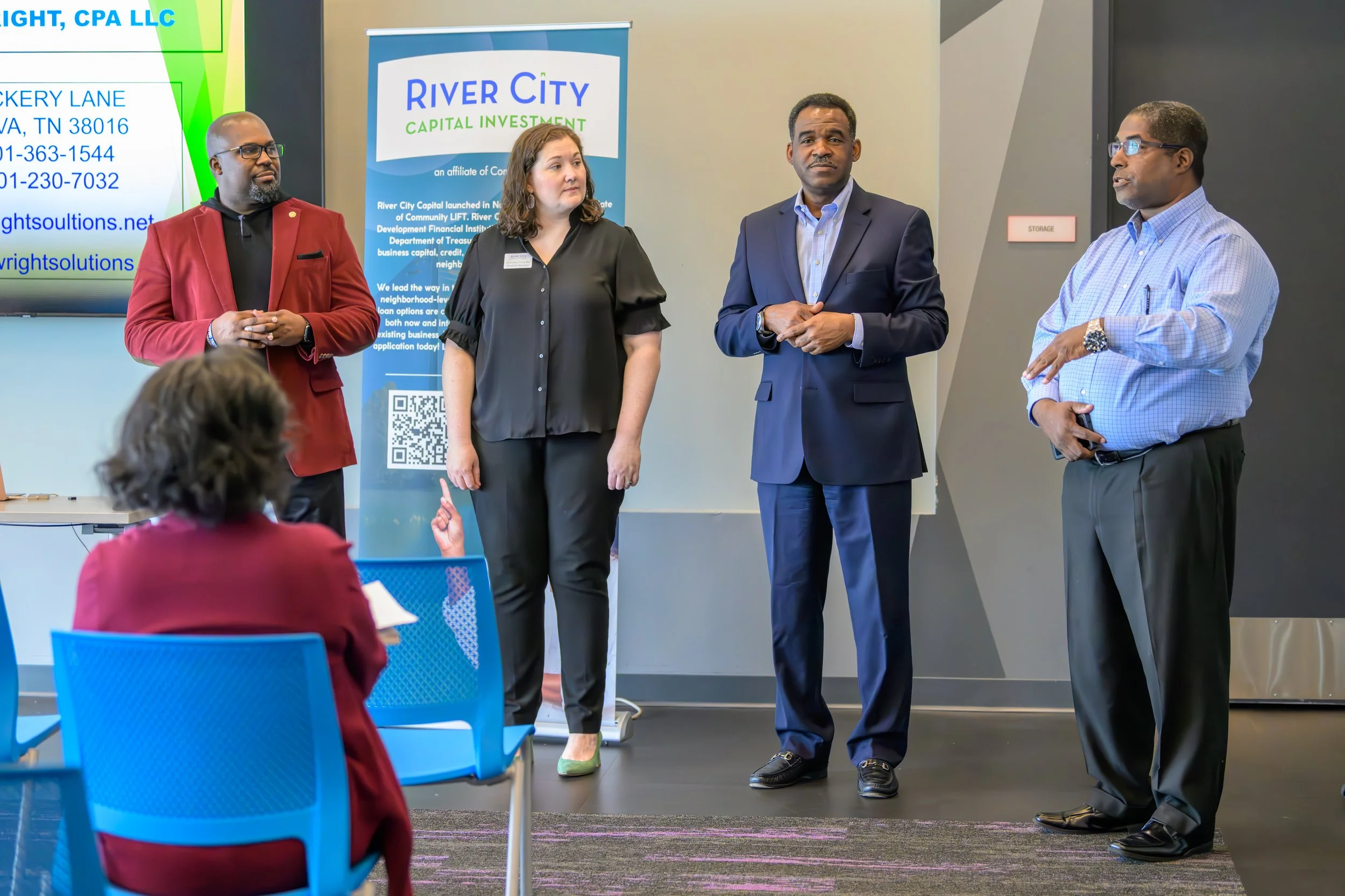Four professionals stand in front of a presentation screen and banner, engaging with an audience in a conference room. They are dressed in business attire, with two men in suits, one woman in a black blouse, and another man in a red blazer. Audience members are seated, including a woman in a pink sweater in the foreground.
