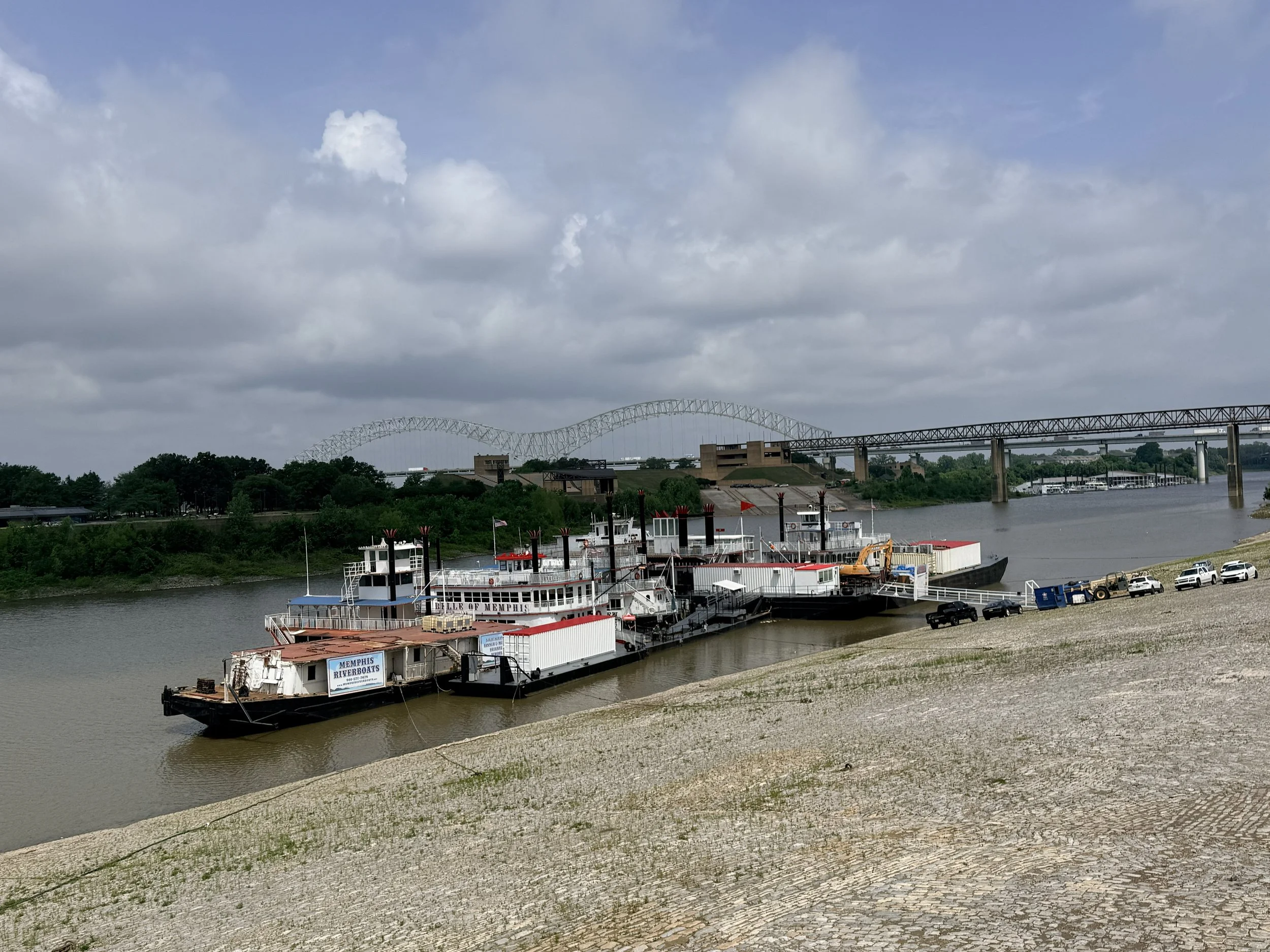View of a river with docked riverboats, a bridge in the background, and cloudy skies overhead.