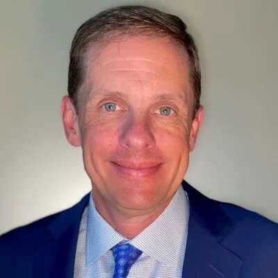 Portrait of a man with short brown hair wearing a navy suit, light blue dress shirt, and blue tie, smiling against a neutral background.