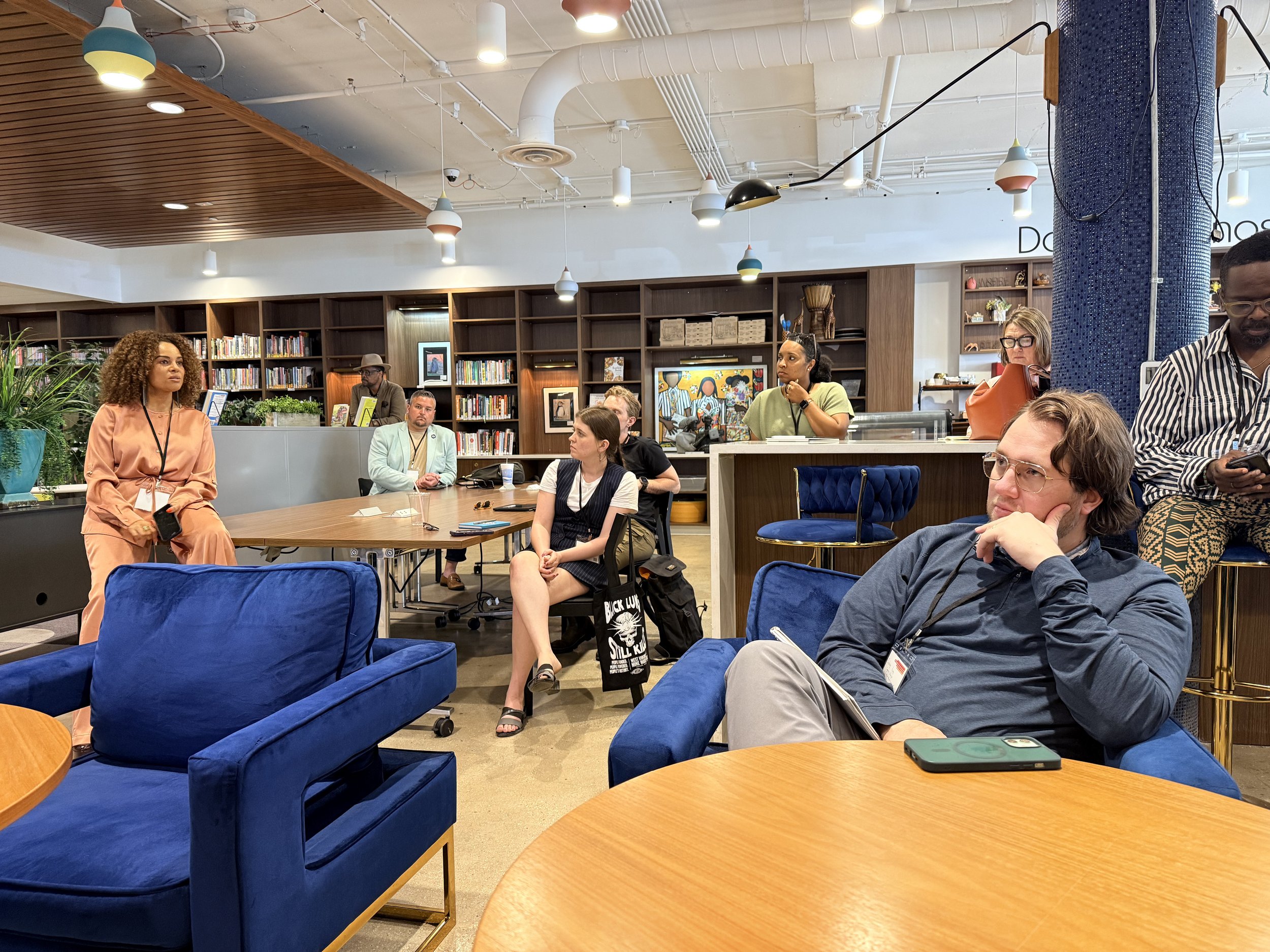 Group of diverse adults sitting and standing in a modern, open-concept library or community space with bookshelves, artwork, and decorative lighting, engaging in a discussion or listening to a speaker.