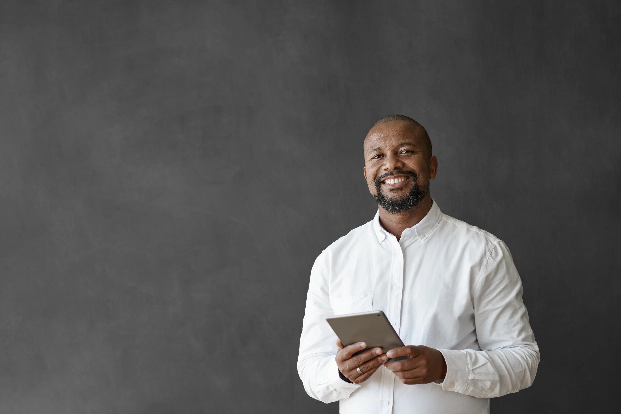 A smiling man with a beard and mustache wearing a white shirt holding a tablet against a dark gray background.