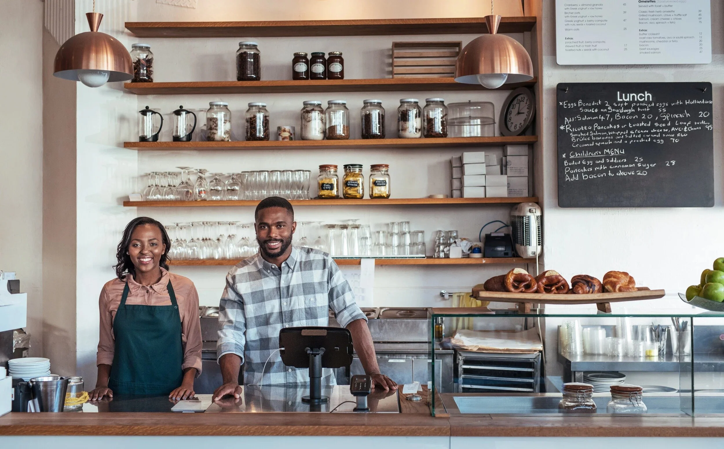 Two staff members, a woman and a man, standing behind the counter of a cafe with shelves holding jars and coffee equipment behind them.