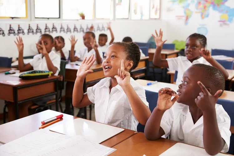 Classroom full of young students in white uniforms raising hands, smiling, and engaging in lesson.