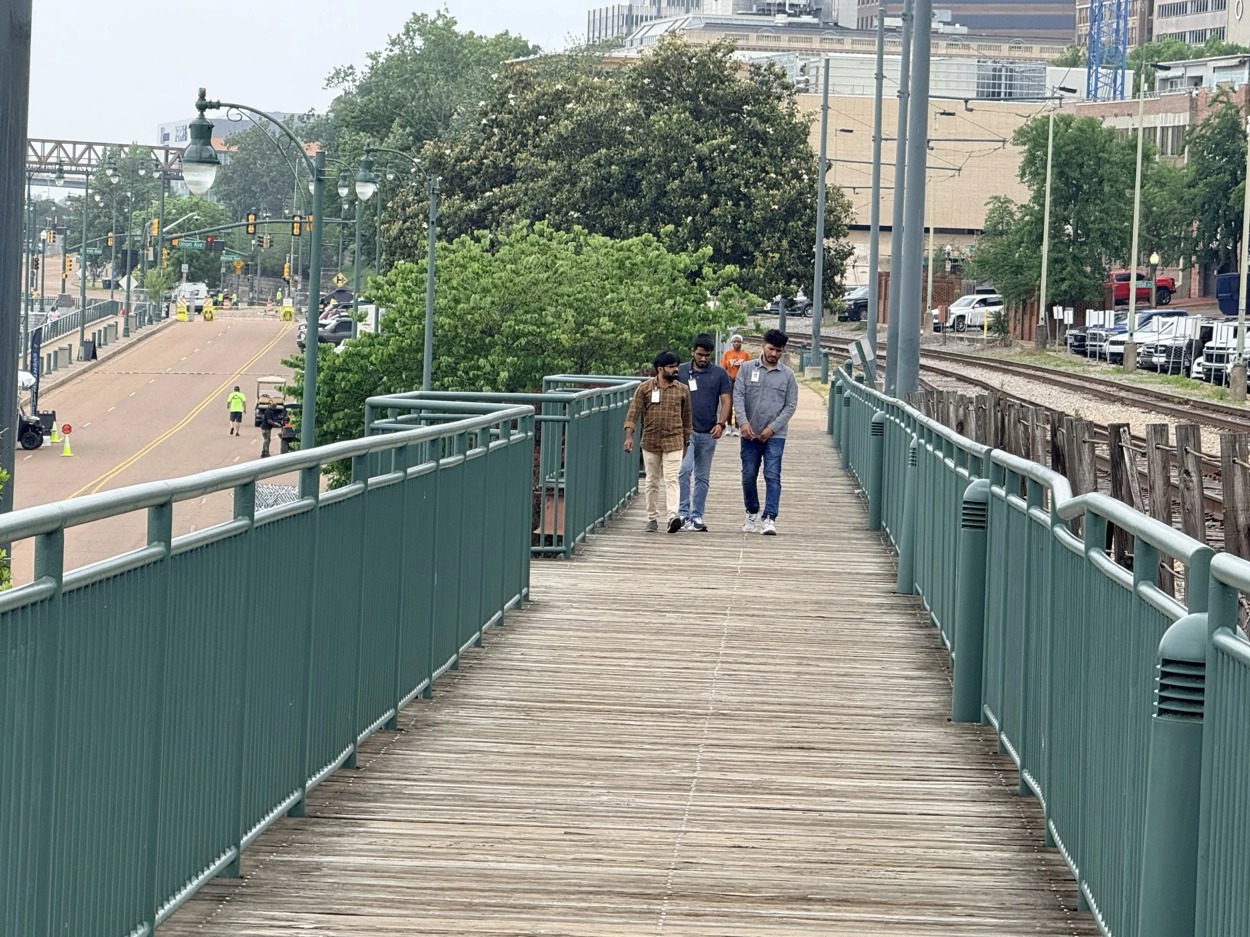 Group of four men walking on a wooden pedestrian bridge next to train tracks in an urban area with trees and buildings.