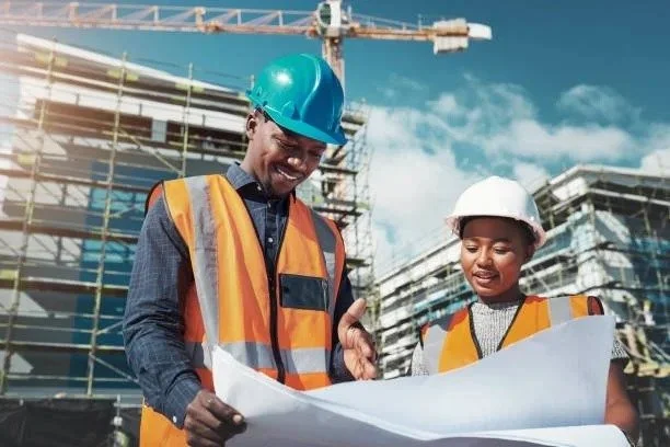 Two construction workers, a man and a woman, wearing safety helmets and orange safety vests, looking at blueprints together at a construction site with a crane and building framing in the background.