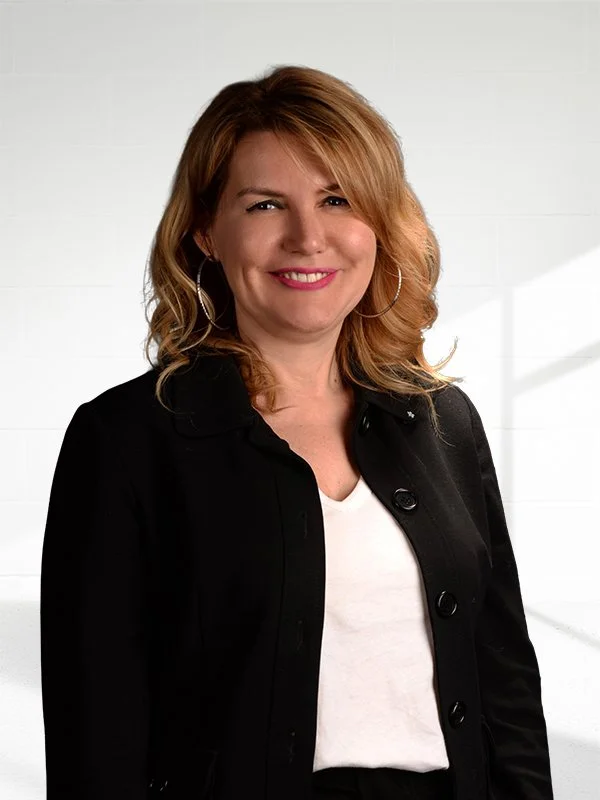 Gülümseyen kadın, siyah ceket ve beyaz tişört giymiş, uzun saçları omuz hizasında, büyük halka küpe takmış.