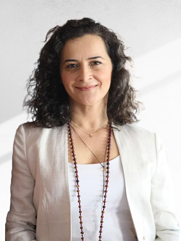 Gülümseyen kadın, beyaz bluz ve ceket giyiyor, uzun boncuklu takı takmış.
