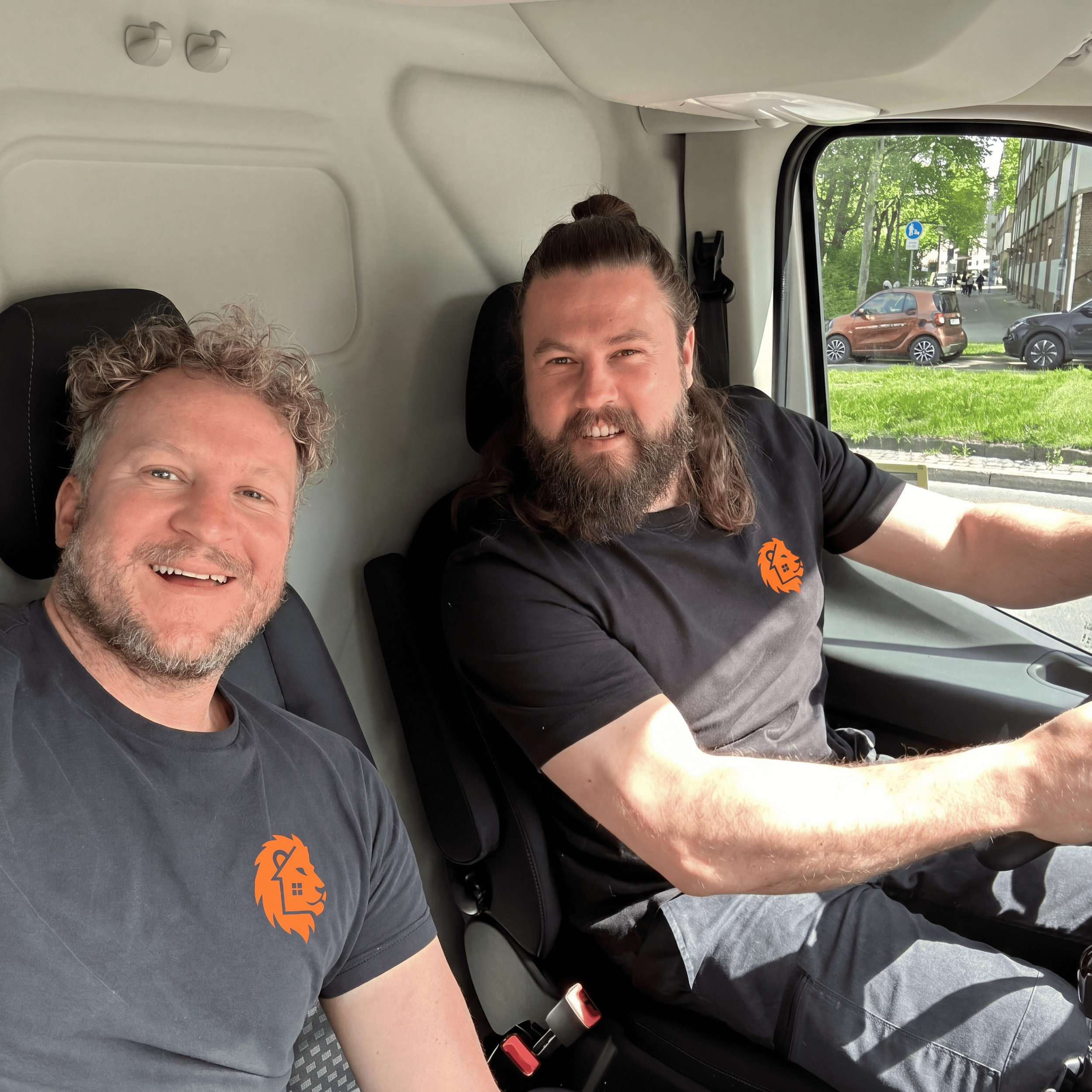 Zwei Männer sitzen nebeneinander im Auto. Der Mann auf der linken Seite hat blondes, lockiges Haar und trägt ein graues T-Shirt mit einem orangefarbenen Löwenlogo. Der Mann auf der rechten Seite hat lange, braune Haare, einen Bart und trägt ein schwarzes T-Shirt. Durch das Autofenster ist eine grüne, städtische Umgebung mit Bäumen, einem blauen Fußgängerhinweisschild, einem braunen und einem schwarzen Auto sichtbar.