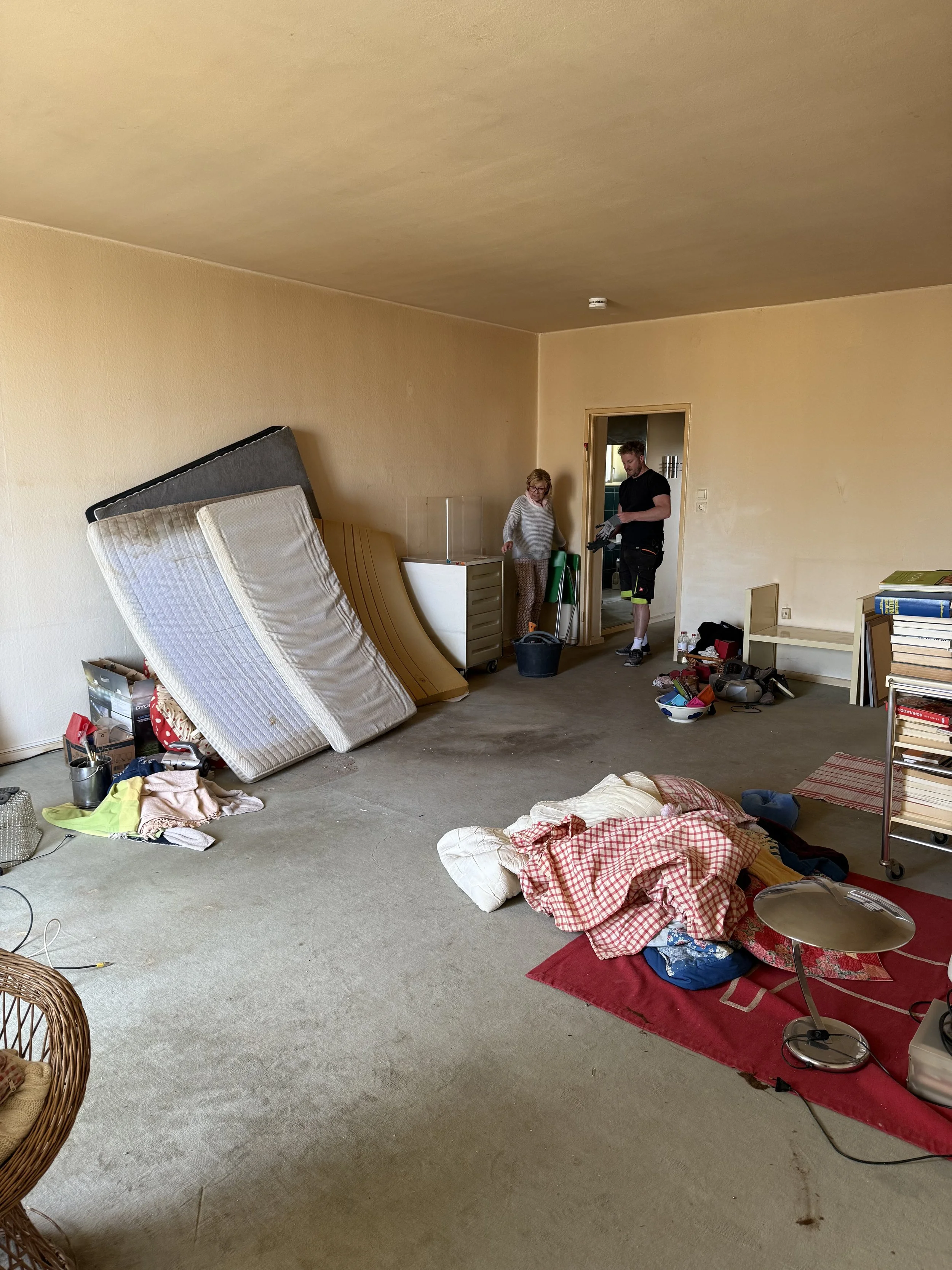 Ein unordentliches Zimmer mit auf dem Boden verstreuten Gegenständen, Bettgestell und Matratze, Teppich, einer Person und einem Hund, die sich im Raum befinden.