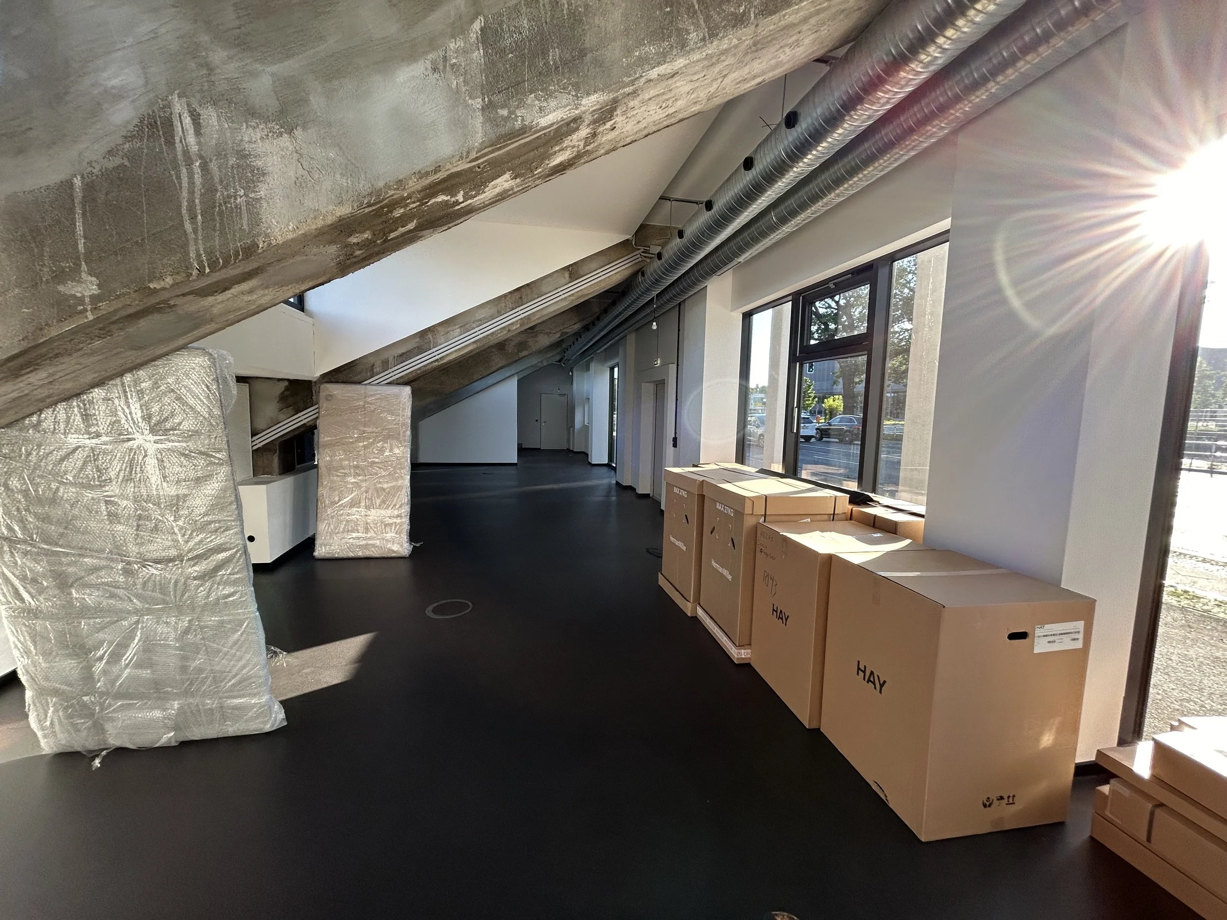 Innenraum eines unvollendeten Gebäudes mit sichtbaren Rohrleitungen und verpackten Baumaterialien, Sonnenlicht strahlt durch große Fenster