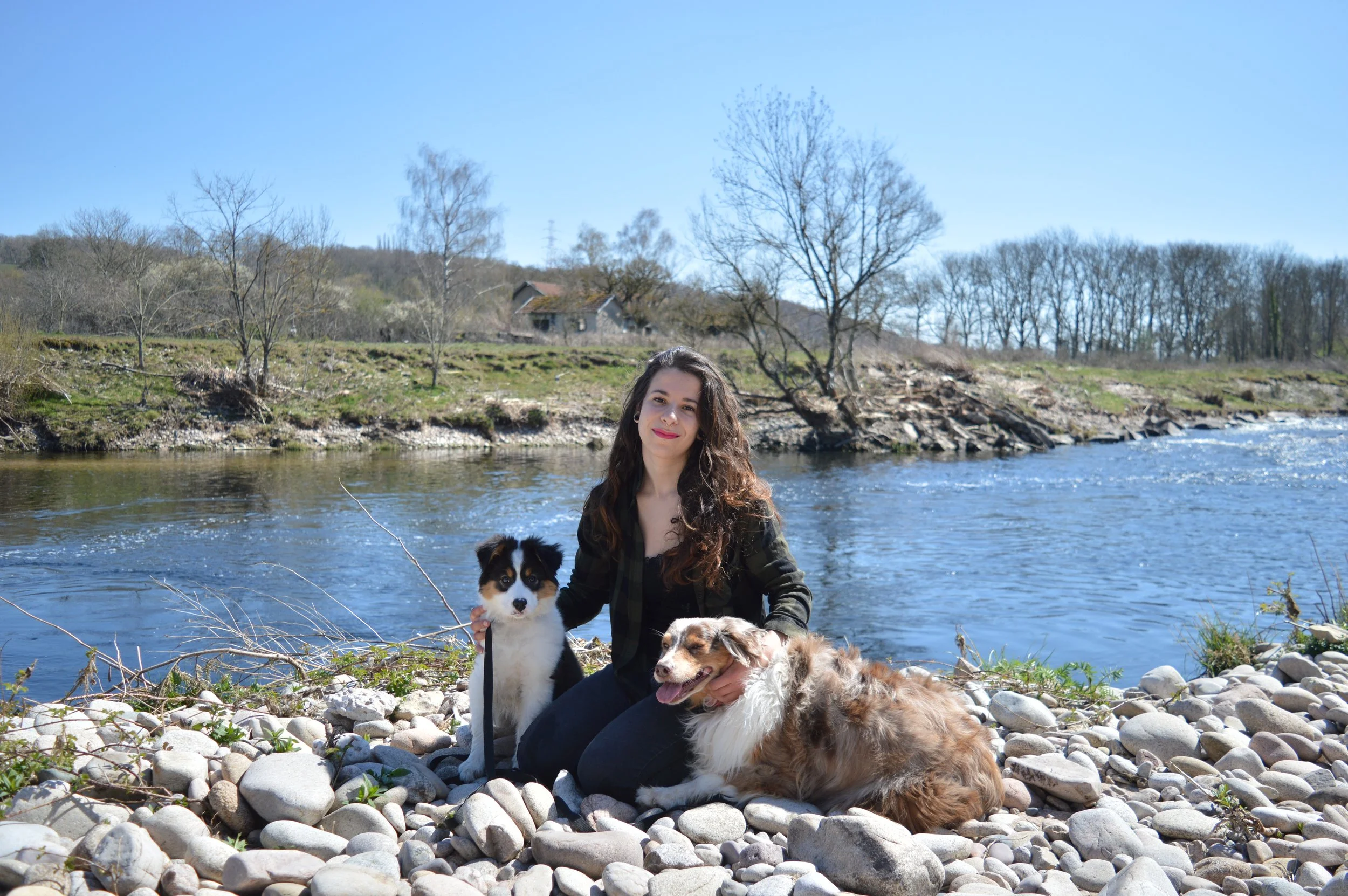 Femme educatrice canin avec un chiot berger australien et un chien berger australien rouge merle devant une rivière dans la nature