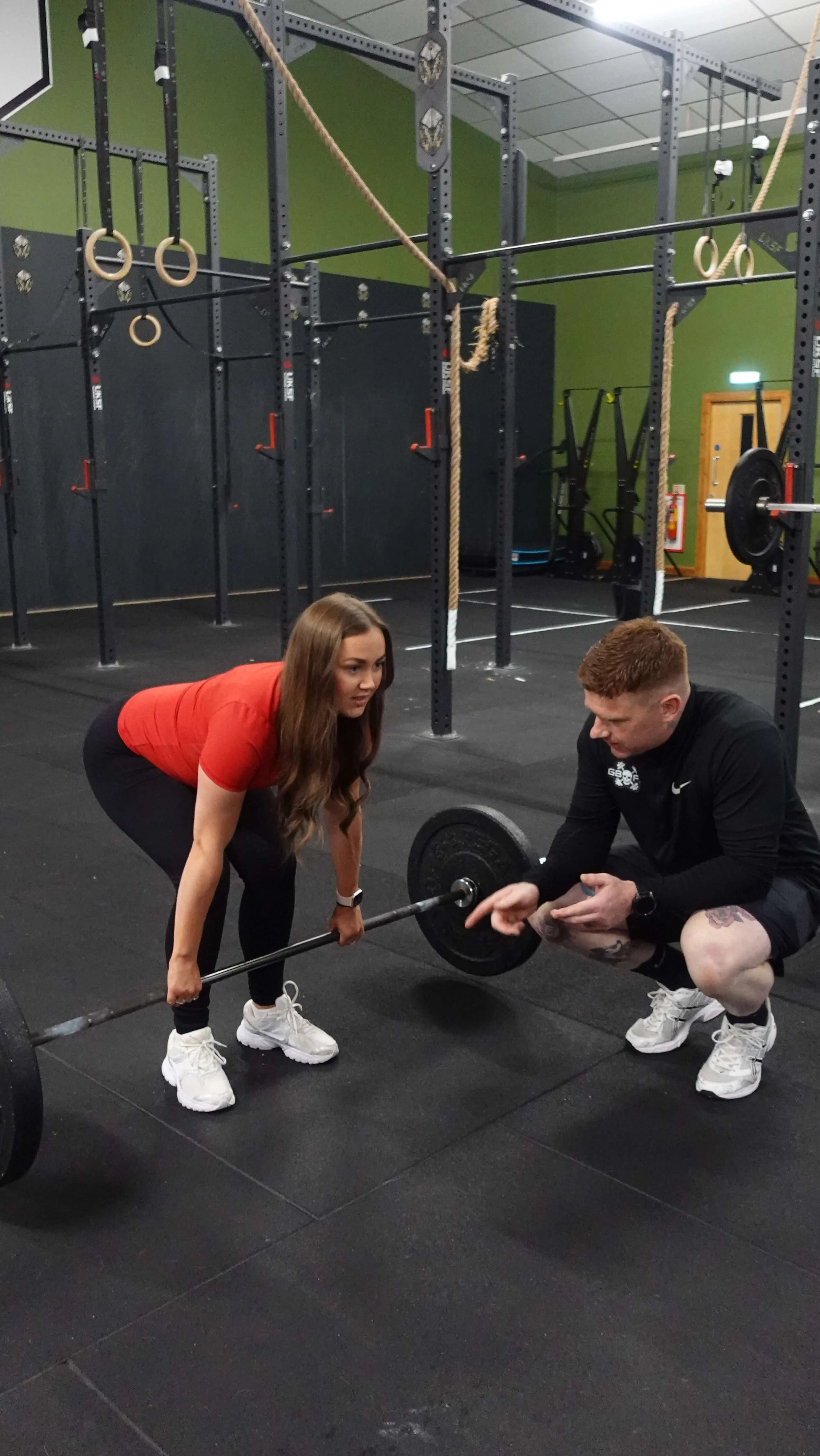 A woman and a man in a gym during a weightlifting workout. The woman is bent over holding a barbell, preparing to lift it, while the man, squatting, shows her proper form or technique. The gym has an industrial look with black flooring, barbells, gymnastic rings, and a green wall.