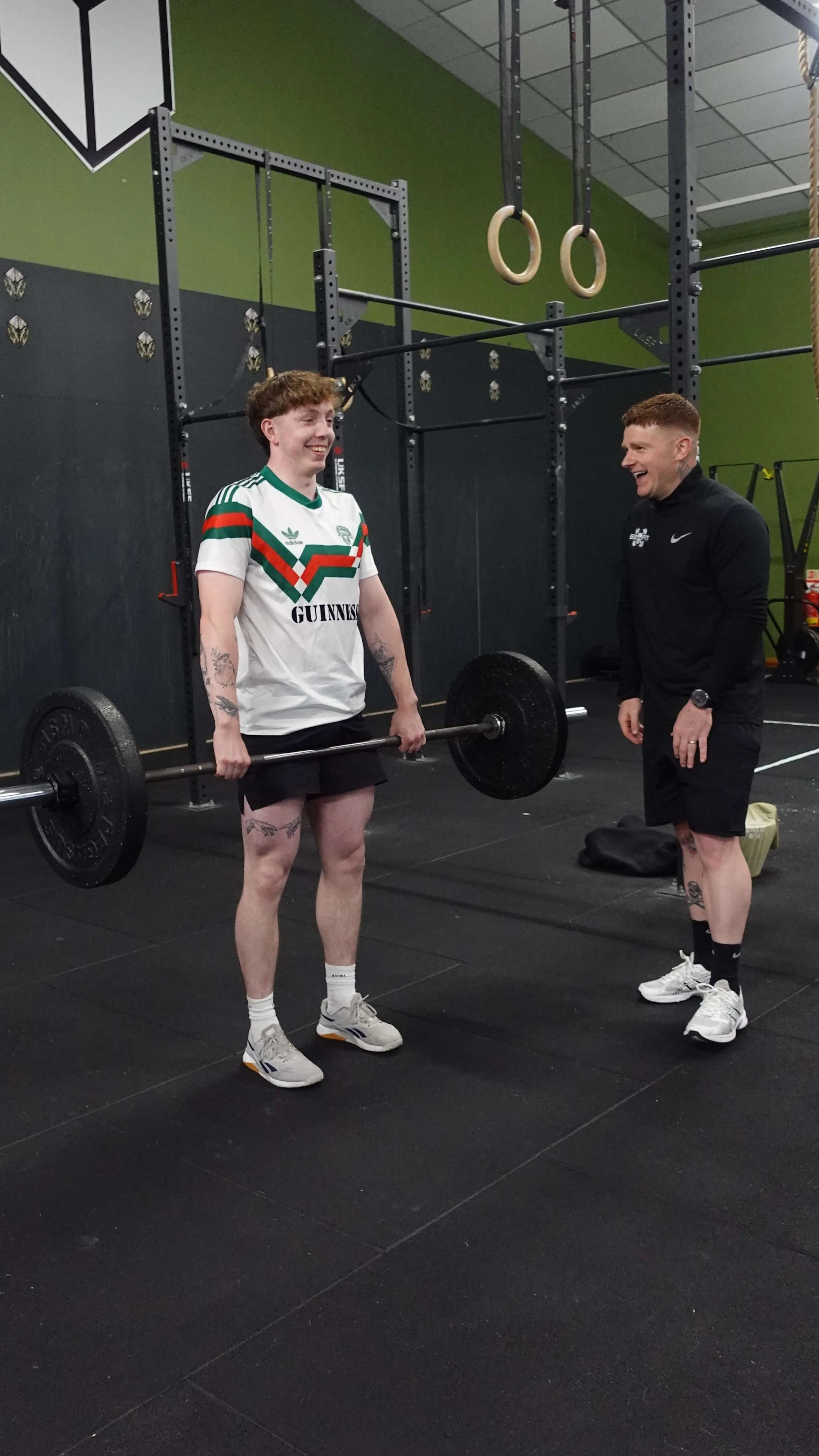 A young man lifting a barbell in a gym, smiling, with a trainer standing nearby, laughing, in front of gym equipment including gymnastic rings.