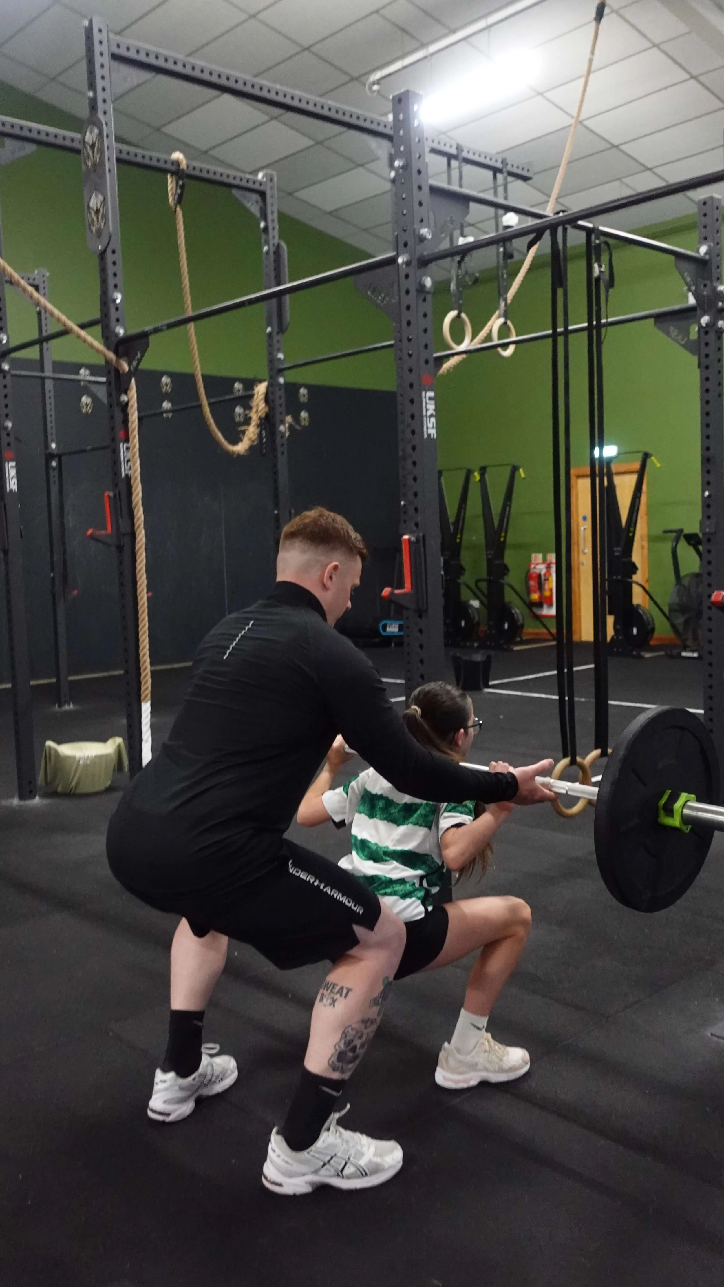 A man supervising a young girl as she performs a squat exercise with a barbell in a gym. The gym has black and green walls, gymnastic rings, ropes, and fitness equipment in the background.