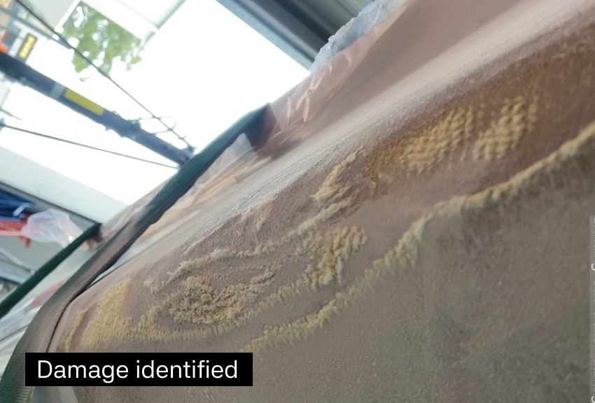 Close-up of a damaged surface, likely a countertop or table, with visible scratches and chipping. In the background, part of a workspace or garage can be seen, including window and some tools.