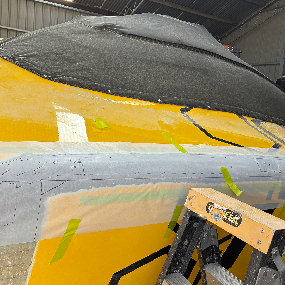 Close-up of a boat with a yellow hull, covered with a black protective cover, inside a storage or workshop with metal walls and ceiling, and a wooden step ladder nearby.