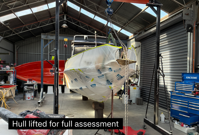 An indoor boat workshop with a boat lifted on blocks for inspection, covered in protective wrapping, with various tools and equipment around.