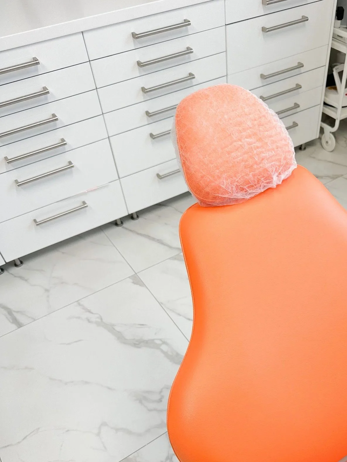 Une chaise orange avec un coussin en tissu bleu clair dans une clinique ou un cabinet médical, avec des tiroirs blancs et un sol en marbre blanc.