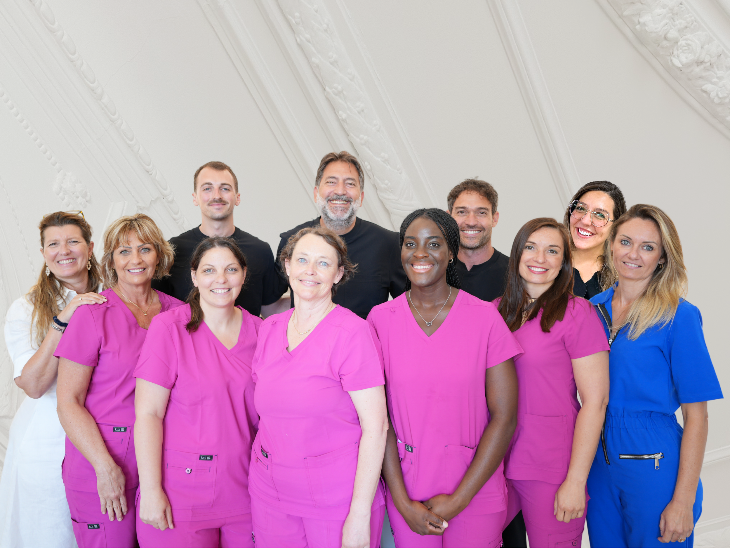 Groupe de professionnels de la santé souriants, portant des blouses ou uniformes colorés, dans une pièce avec un plafond orné de moulures décoratives.