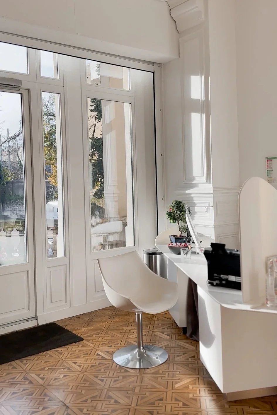 Coin d'accueil avec une porte-fenêtre, une chaise blanche, un bureau blanc avec une petite plante et des objets divers, parquet en bois, intérieur lumineux.
