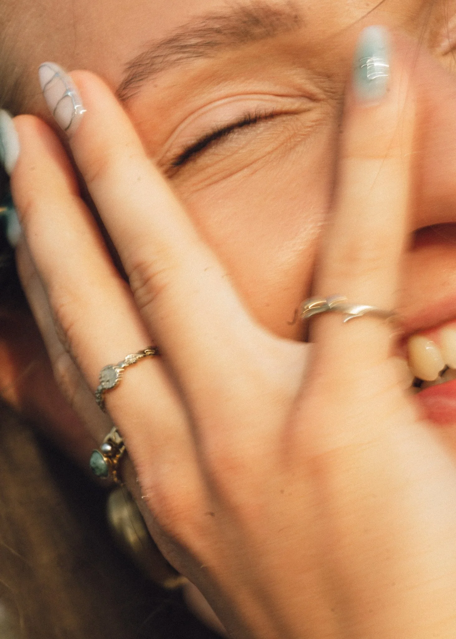 Close-up of a person's face with closed eyes, hand with rings near face, and nail art with silver and turquoise accents.