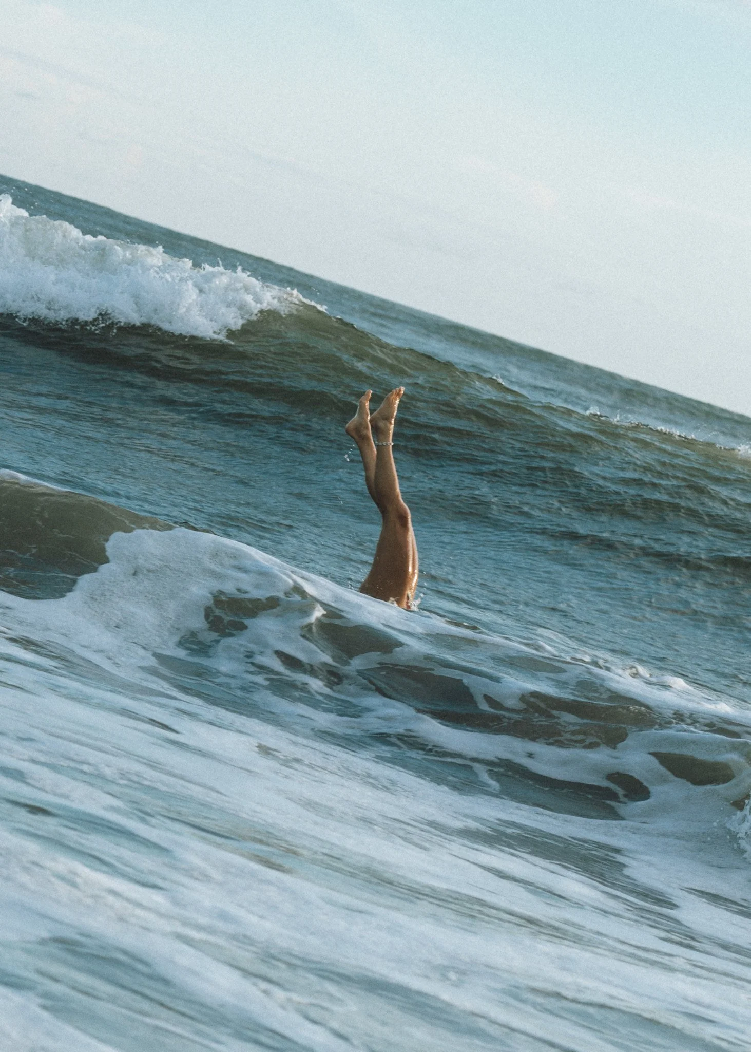 Person swimming in the ocean with legs and feet visible above the water, a wave approaching, and a clear sky.