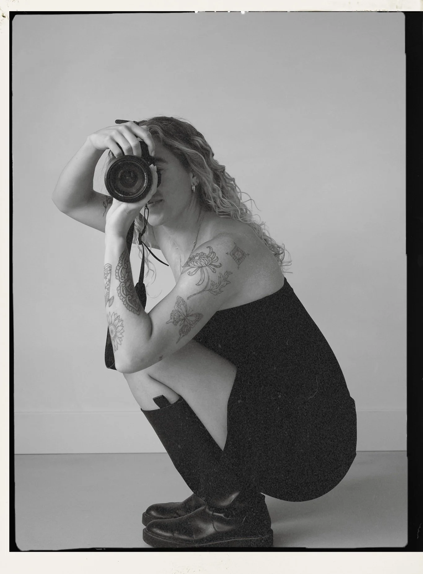 A woman with curly hair taking a mirror selfie with a camera, crouching down with her left arm resting on her knee, tattoos on her arms and shoulders, wearing a black backless top, black pants, and black shoes, against a plain light-colored wall.